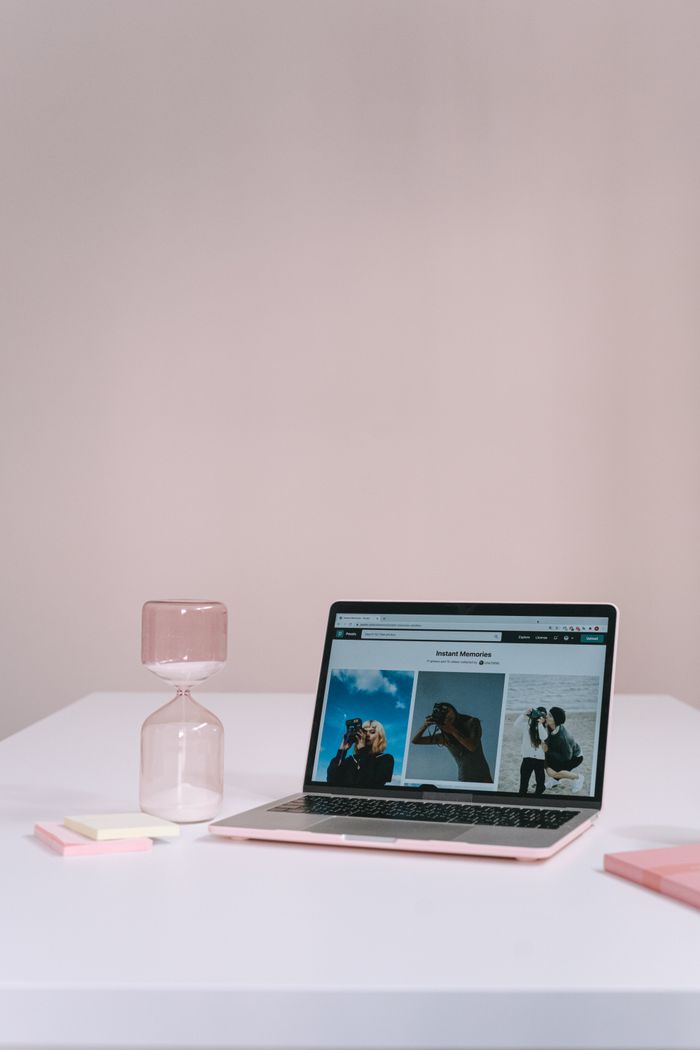 A clean, minimalist workspace featuring a laptop with images on the screen and a pink glass hourglass on a white table.